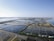 Aerial view of salt evaporation ponds located near a coastal area. The ponds form a grid-like pattern with various sections of different colors, ranging from dark blues to light browns and yellows. The horizon is clear and sky is blue with a few distant structures visible along the shoreline.