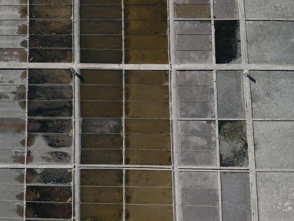 An aerial view of a grid-like pattern consisting of rectangular sections filled with different earthy tones, possibly representing salt pans or agricultural plots. The sections vary in color from dark brown to light gray, with divider lines creating a structured layout.