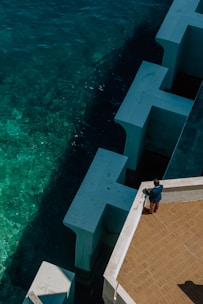 A person stands on a terracotta-tiled terrace next to large white angular structures bordering deep blue-green ocean water. The scene captures a bird's-eye view, emphasizing the contrast between the geometric forms and organic movement of the sea.