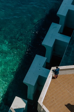 A person stands on a terracotta-tiled terrace next to large white angular structures bordering deep blue-green ocean water. The scene captures a bird's-eye view, emphasizing the contrast between the geometric forms and organic movement of the sea.