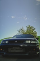 A black sports car is prominently displayed against a backdrop of a blue sky and some greenery. The car is positioned in the foreground, exhibiting its sleek and polished exterior with a focus on the front grille and headlights. A hint of sunlight reflects subtly on its surface, enhancing its glossy finish.