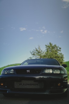 A black sports car is prominently displayed against a backdrop of a blue sky and some greenery. The car is positioned in the foreground, exhibiting its sleek and polished exterior with a focus on the front grille and headlights. A hint of sunlight reflects subtly on its surface, enhancing its glossy finish.