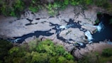 An aerial view of a river flowing through a rocky landscape, surrounded by lush green trees. The river cuts through large rock formations with visible rapids and small waterfalls. The contrast between the dark water and light-colored rocks creates a striking visual.