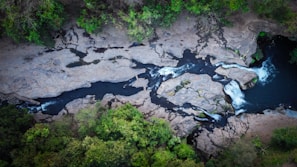 An aerial view of a river flowing through a rocky landscape, surrounded by lush green trees. The river cuts through large rock formations with visible rapids and small waterfalls. The contrast between the dark water and light-colored rocks creates a striking visual.