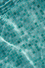 A close-up view of a tiled surface, consisting of small, square tiles in various shades of turquoise and blue. The light reflecting off the water creates a wavy, shimmering effect over the tiles.