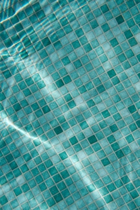 A close-up view of a tiled surface, consisting of small, square tiles in various shades of turquoise and blue. The light reflecting off the water creates a wavy, shimmering effect over the tiles.