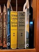 A collection of classic books on a wooden shelf, featuring titles by the author Maurice Leblanc. Prominently displayed are editions of 'Arsène Lupin Versus Herlock Sholmes' with distinctive yellow and olive covers, published by J.S. Ogilvie Publishing Company. Other featured titles include 'The Hollow Needle' and 'The Blonde Phantom.' Some book spines have decorative elements and labels are attached within the books.