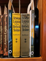 A collection of classic books on a wooden shelf, featuring titles by the author Maurice Leblanc. Prominently displayed are editions of 'Arsène Lupin Versus Herlock Sholmes' with distinctive yellow and olive covers, published by J.S. Ogilvie Publishing Company. Other featured titles include 'The Hollow Needle' and 'The Blonde Phantom.' Some book spines have decorative elements and labels are attached within the books.