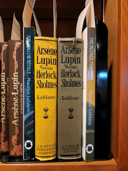 A collection of classic books on a wooden shelf, featuring titles by the author Maurice Leblanc. Prominently displayed are editions of 'Arsène Lupin Versus Herlock Sholmes' with distinctive yellow and olive covers, published by J.S. Ogilvie Publishing Company. Other featured titles include 'The Hollow Needle' and 'The Blonde Phantom.' Some book spines have decorative elements and labels are attached within the books.