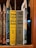 A collection of classic books on a wooden shelf, featuring titles by the author Maurice Leblanc. Prominently displayed are editions of 'Arsène Lupin Versus Herlock Sholmes' with distinctive yellow and olive covers, published by J.S. Ogilvie Publishing Company. Other featured titles include 'The Hollow Needle' and 'The Blonde Phantom.' Some book spines have decorative elements and labels are attached within the books.