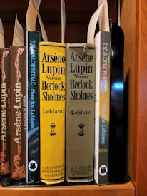 A collection of classic books on a wooden shelf, featuring titles by the author Maurice Leblanc. Prominently displayed are editions of 'Arsène Lupin Versus Herlock Sholmes' with distinctive yellow and olive covers, published by J.S. Ogilvie Publishing Company. Other featured titles include 'The Hollow Needle' and 'The Blonde Phantom.' Some book spines have decorative elements and labels are attached within the books.