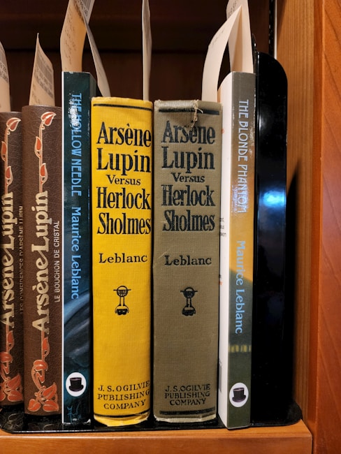 A collection of classic books on a wooden shelf, featuring titles by the author Maurice Leblanc. Prominently displayed are editions of 'Arsène Lupin Versus Herlock Sholmes' with distinctive yellow and olive covers, published by J.S. Ogilvie Publishing Company. Other featured titles include 'The Hollow Needle' and 'The Blonde Phantom.' Some book spines have decorative elements and labels are attached within the books.