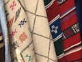 Multiple pieces of fabric with various woven patterns. The fabrics feature intricate designs, including geometric shapes, lines, and crosses. The colors range from neutral tones like beige and white to vibrant hues such as red, blue, and green.