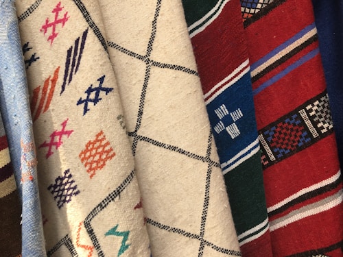 Multiple pieces of fabric with various woven patterns. The fabrics feature intricate designs, including geometric shapes, lines, and crosses. The colors range from neutral tones like beige and white to vibrant hues such as red, blue, and green.