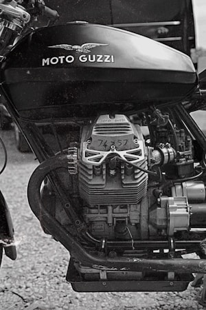 A close-up view of a vintage motorcycle engine with the Moto Guzzi logo prominently displayed on the fuel tank. The image is in black and white, showcasing the mechanical details and textures of the engine components.