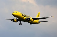 A brightly colored yellow airplane is flying against a cloudy sky. The aircraft has the brand name prominently displayed on its fuselage and tail. The landing gear is visible as it is lowered, suggesting the plane is in the process of either landing or taking off.