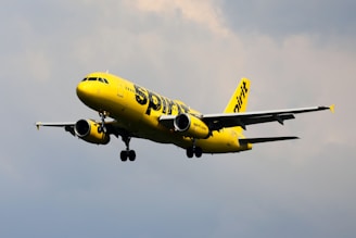 A brightly colored yellow airplane is flying against a cloudy sky. The aircraft has the brand name prominently displayed on its fuselage and tail. The landing gear is visible as it is lowered, suggesting the plane is in the process of either landing or taking off.