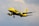 A brightly colored yellow airplane is flying against a cloudy sky. The aircraft has the brand name prominently displayed on its fuselage and tail. The landing gear is visible as it is lowered, suggesting the plane is in the process of either landing or taking off.