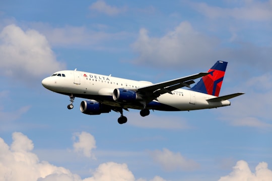 A passenger airplane in flight against a background of blue sky and scattered clouds. The aircraft is white with blue and red accents and features prominent jet engines beneath the wings.