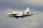 A sleek, modern private jet is flying against a cloudy sky. The aircraft is white with a streamlined design and dark accents on its tail and wings.