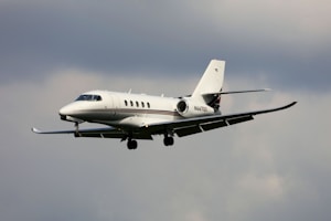 A sleek, modern private jet is flying against a cloudy sky. The aircraft is white with a streamlined design and dark accents on its tail and wings.