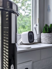 A white security camera is placed on a windowsill, alongside a small decorative plant in a geometric-patterned pot. The window provides a view of green foliage outside, suggesting a natural setting. The area appears to be inside a house, and the grey tones create a modern vibe.