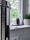 A white security camera is placed on a windowsill, alongside a small decorative plant in a geometric-patterned pot. The window provides a view of green foliage outside, suggesting a natural setting. The area appears to be inside a house, and the grey tones create a modern vibe.