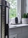 A white security camera is placed on a windowsill, alongside a small decorative plant in a geometric-patterned pot. The window provides a view of green foliage outside, suggesting a natural setting. The area appears to be inside a house, and the grey tones create a modern vibe.