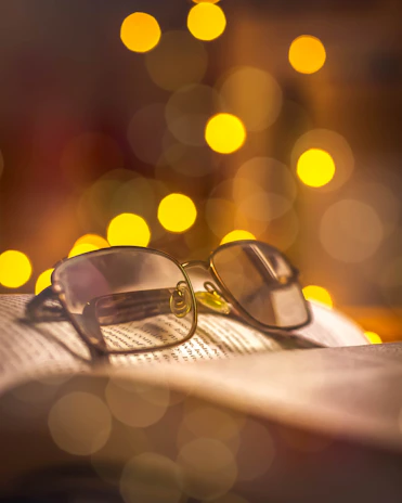 A pair of eyeglasses rests on an open book with pages visible, with a warm bokeh effect of yellow lights in the background adding a cozy ambiance.