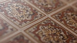 An intricate pattern of ceramic tiles with a vintage design. The tiles feature symmetrical floral motifs with a central rosette surrounded by swirling patterns. The color scheme includes earthy tones of brown, beige, and cream, emphasizing the classic and ornate style.