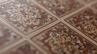 An intricate pattern of ceramic tiles with a vintage design. The tiles feature symmetrical floral motifs with a central rosette surrounded by swirling patterns. The color scheme includes earthy tones of brown, beige, and cream, emphasizing the classic and ornate style.