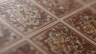 An intricate pattern of ceramic tiles with a vintage design. The tiles feature symmetrical floral motifs with a central rosette surrounded by swirling patterns. The color scheme includes earthy tones of brown, beige, and cream, emphasizing the classic and ornate style.
