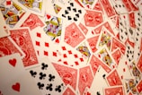 A vibrant assortment of playing cards scattered in a chaotic manner. The cards display various suits and numbers, predominantly featuring the colors red and black with intricate designs. Several face cards are visible, showcasing traditional royal characters in vivid detail.