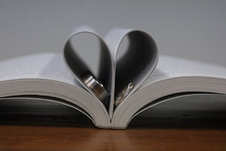 An open book with pages folded inward to create the shape of a heart, containing a couple of rings inside the fold. The book rests on a wooden surface.
