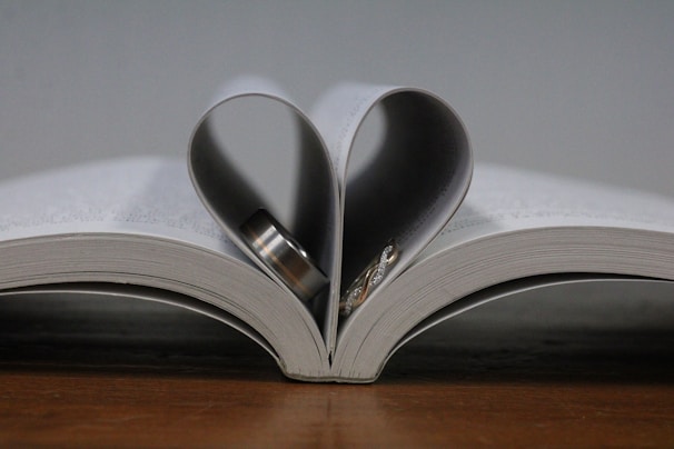 An open book with pages folded inward to create the shape of a heart, containing a couple of rings inside the fold. The book rests on a wooden surface.
