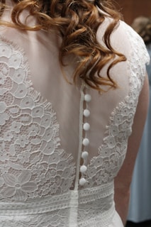Intricate lace details with floral patterns adorn the back of a white garment, featuring a row of small white buttons. The fabric appears delicate and elegant, complemented by wavy hair cascading over the shoulders.