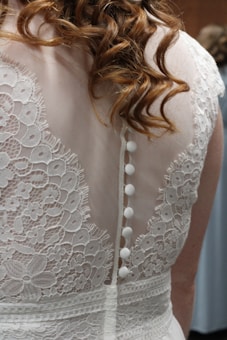Intricate lace details with floral patterns adorn the back of a white garment, featuring a row of small white buttons. The fabric appears delicate and elegant, complemented by wavy hair cascading over the shoulders.
