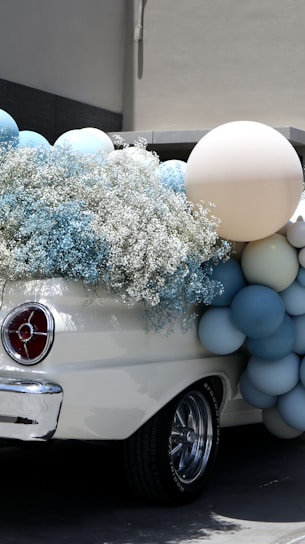 An elegant vintage car is adorned with pastel-colored balloons and delicate white flowers, creating a festive and charming atmosphere.