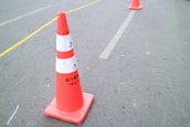 A bright orange traffic cone with reflective white bands is placed on a paved road. Another traffic cone is visible in the background, and a thin yellow line runs diagonally across the image. The road surface appears slightly textured.