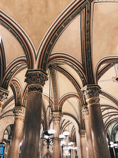 Elegant, high-arched ceilings supported by intricately decorated columns in a historic building interior. Ornate designs and detailed craftsmanship adorn the architecture, with hanging light fixtures adding to the majestic ambiance.