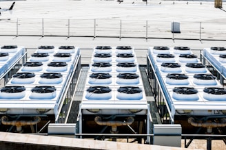 Rows of industrial air conditioning units are arranged on a rooftop, each featuring large fans housed in square metal enclosures. The units are aligned in a grid pattern, set against a background of a fenced area with an expansive flat surface.