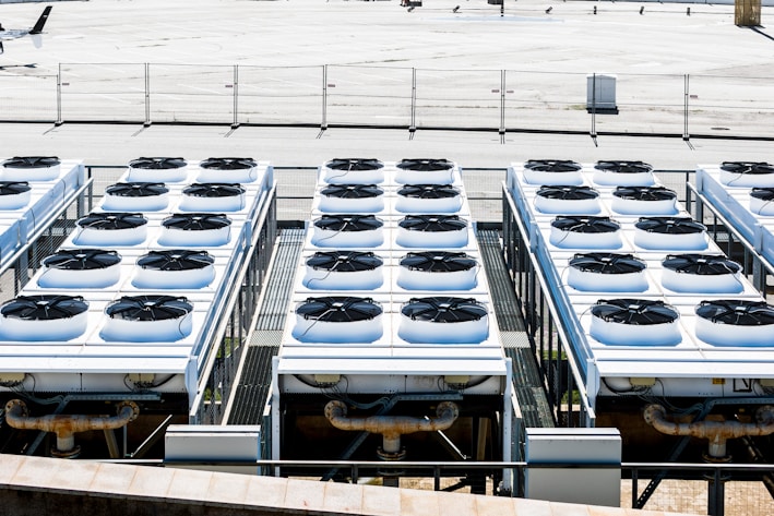 Rows of industrial air conditioning units are arranged on a rooftop, each featuring large fans housed in square metal enclosures. The units are aligned in a grid pattern, set against a background of a fenced area with an expansive flat surface.