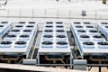Rows of industrial air conditioning units are arranged on a rooftop, each featuring large fans housed in square metal enclosures. The units are aligned in a grid pattern, set against a background of a fenced area with an expansive flat surface.