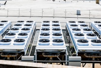 Rows of industrial air conditioning units are arranged on a rooftop, each featuring large fans housed in square metal enclosures. The units are aligned in a grid pattern, set against a background of a fenced area with an expansive flat surface.