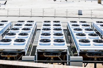 Rows of industrial air conditioning units are arranged on a rooftop, each featuring large fans housed in square metal enclosures. The units are aligned in a grid pattern, set against a background of a fenced area with an expansive flat surface.