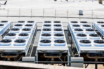 Rows of industrial air conditioning units are arranged on a rooftop, each featuring large fans housed in square metal enclosures. The units are aligned in a grid pattern, set against a background of a fenced area with an expansive flat surface.