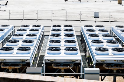 Rows of industrial air conditioning units are arranged on a rooftop, each featuring large fans housed in square metal enclosures. The units are aligned in a grid pattern, set against a background of a fenced area with an expansive flat surface.