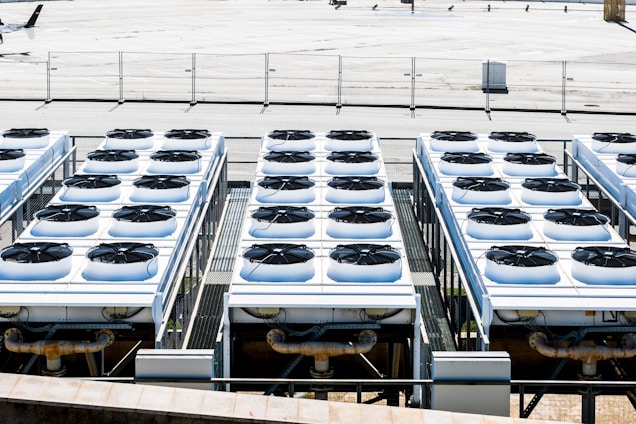 Rows of industrial air conditioning units are arranged on a rooftop, each featuring large fans housed in square metal enclosures. The units are aligned in a grid pattern, set against a background of a fenced area with an expansive flat surface.