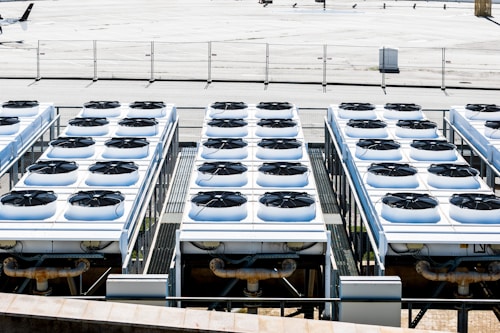Rows of industrial air conditioning units are arranged on a rooftop, each featuring large fans housed in square metal enclosures. The units are aligned in a grid pattern, set against a background of a fenced area with an expansive flat surface.