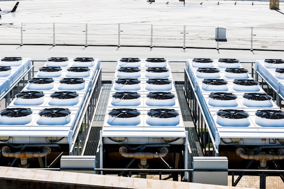 Rows of industrial air conditioning units are arranged on a rooftop, each featuring large fans housed in square metal enclosures. The units are aligned in a grid pattern, set against a background of a fenced area with an expansive flat surface.